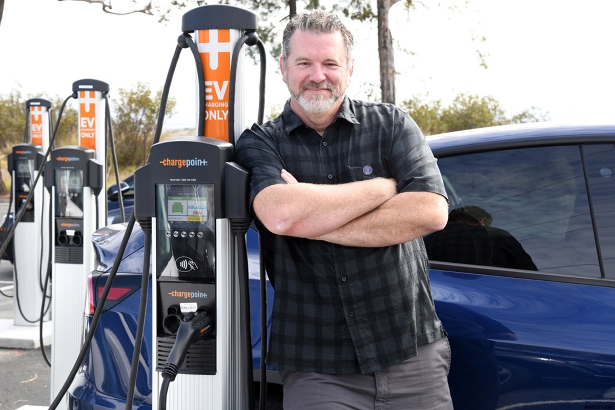A older white gentleman standing with arms crossed next to an electric vehicle charger station.