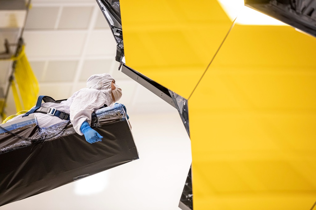 A person in a clean suit lays on a crane to access the James Webb Space Telescope