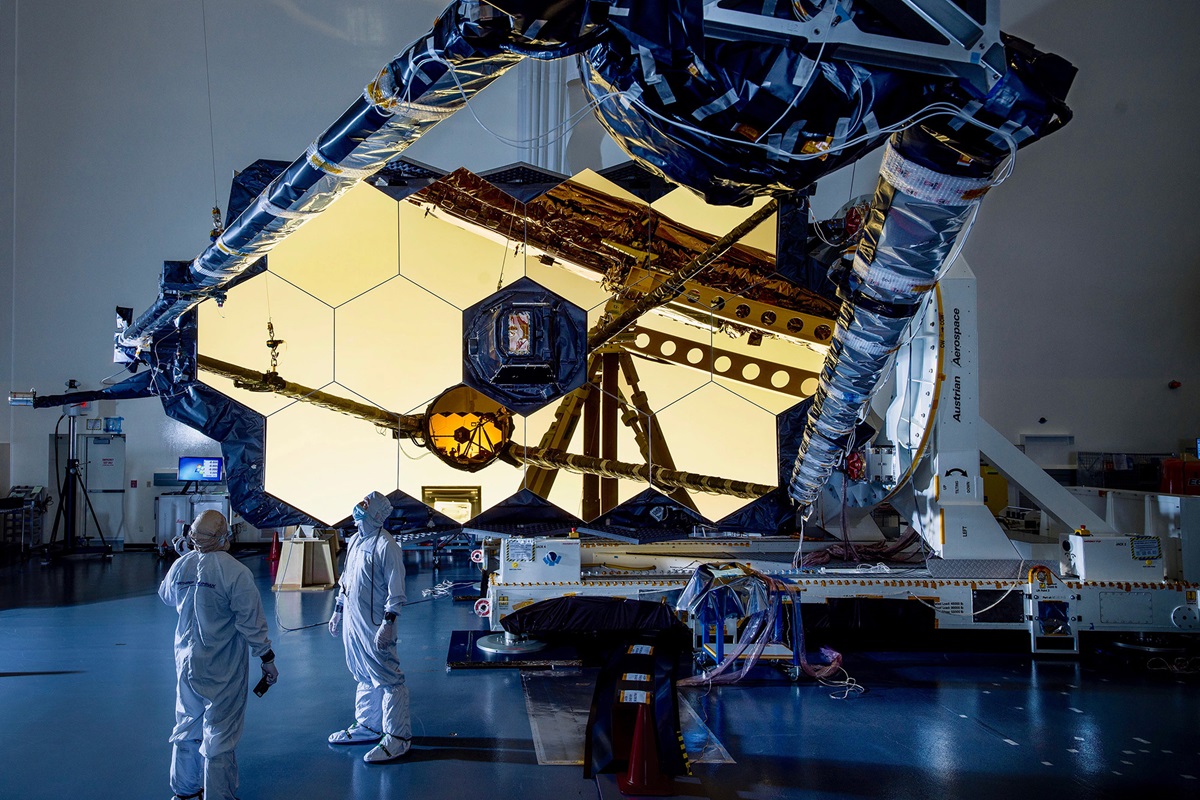 Two people in clean suits look up at the James Webb Space Telescope