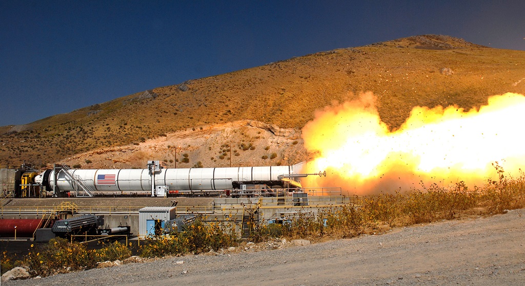 Horizontal rocket moter in desert environment during test fire, with smoke and flames coming out of the back of rocket.