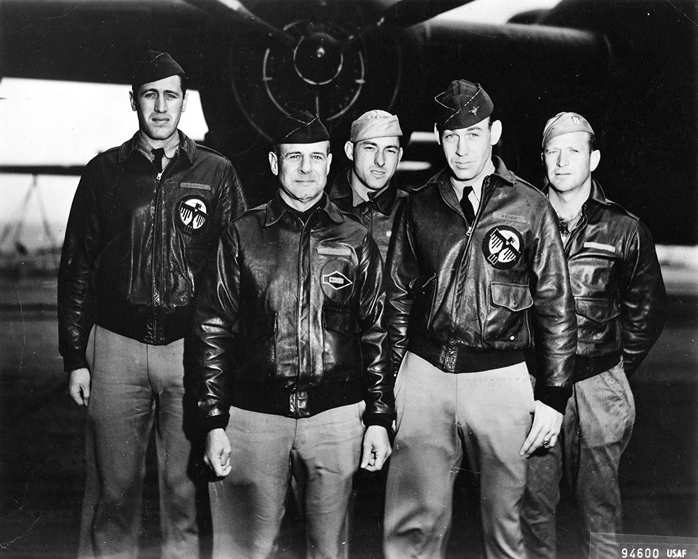 five men in leather coats in front of military plane