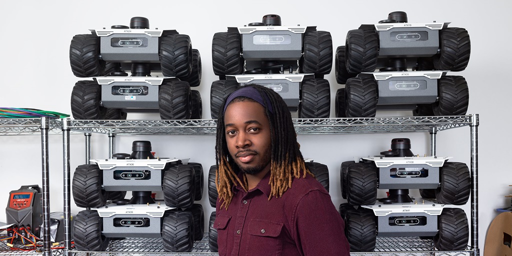 A black man stands in front of a wall of robotic vehicles and looks at the camera