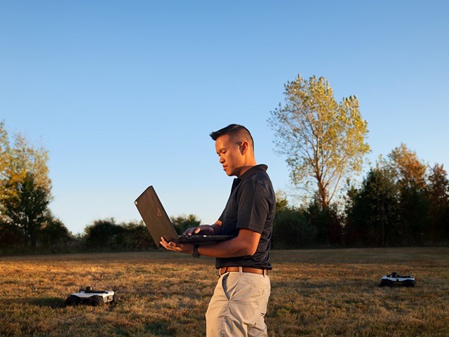A man works on a laptop in a field with robotic vehicles in the background