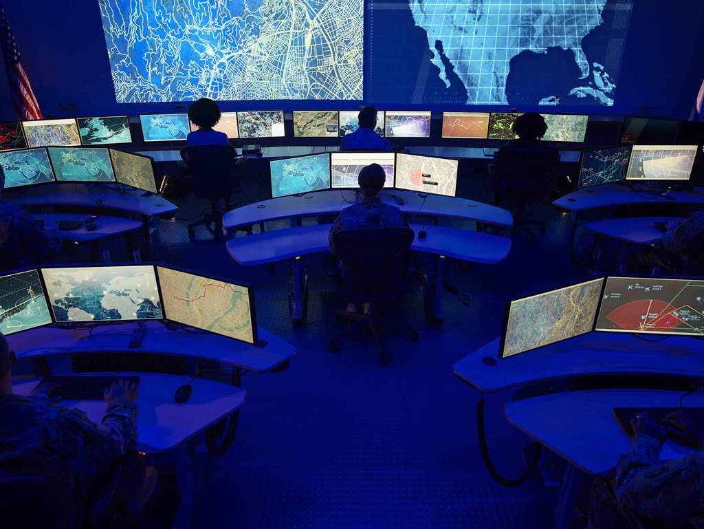 Large command center with multiple computer screens and people monitoring data.