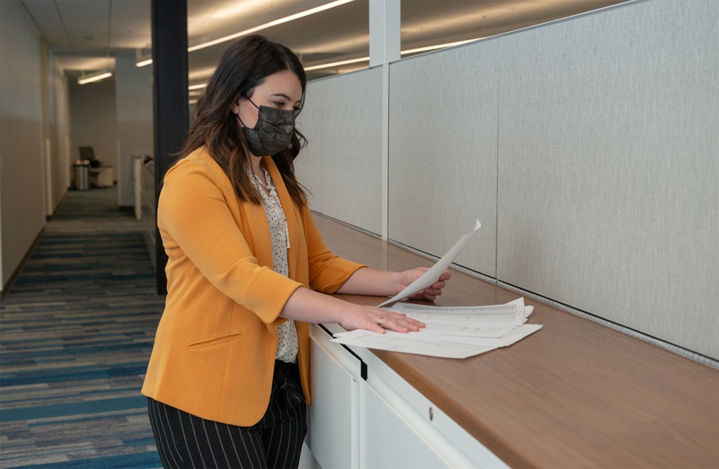 A white woman with a face mask and wearing yellow inside of an office building is standing and looking at papers on a desk