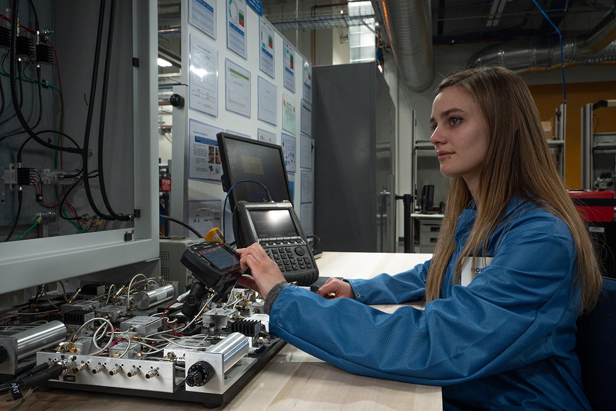 A white female in a blue lab coat in a manufacturing plant operating systems at her desk