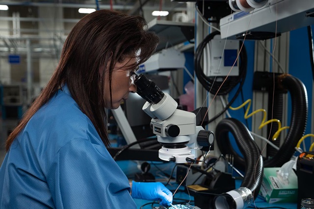 A female person of color in a blue lab coat looking through a telescope in a manufactuing plant