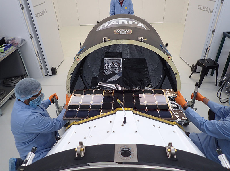 three workers in blue lab uniforms working on spacecraft