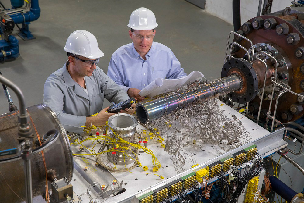 Two men in hard hats look at instruments on a table in a manufacturing setting.