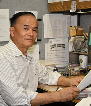 Male sitting at desk with papers in hands smiles for photo