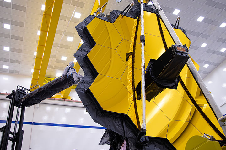 an engineer wearing a clean suit sits on a crane to work on the James Webb Space Telescope.