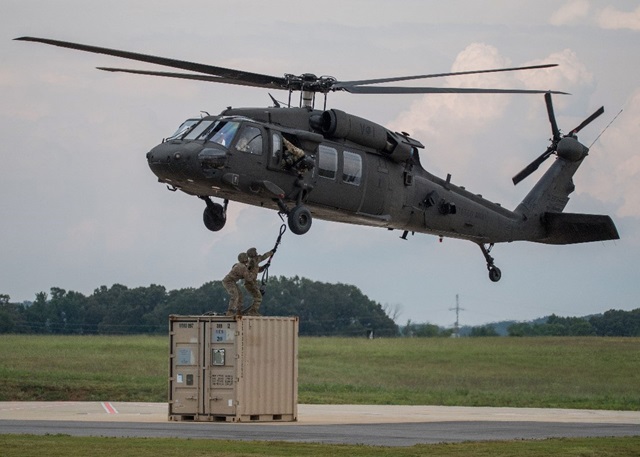 A UH-60V helicopter hovers above two men standing on top of a cargo crate