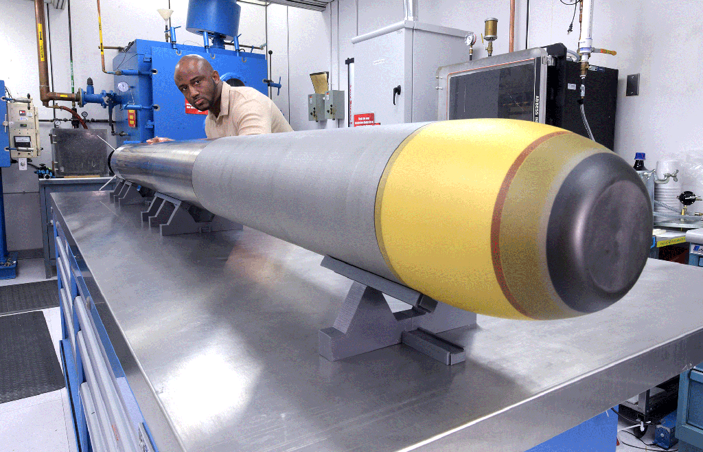 a black man stands next to a Very Lightweight Torpedo (VLWT) in a lab