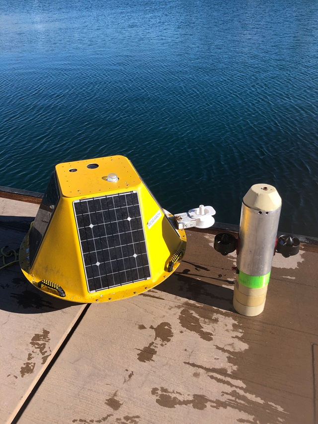 A yellow, eight-sides yellow shape with solar panels is a floating buoy, which sits next to a metal cylinder with small propellors on it. The cylinder holds the sensor package that will be attached to the buoy. The buoy and cylinder sit on a wooden dock with calm water behind them.