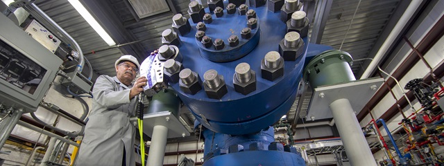Man in lab coat works on largeb blue cylinder in a manufacturing setting.
