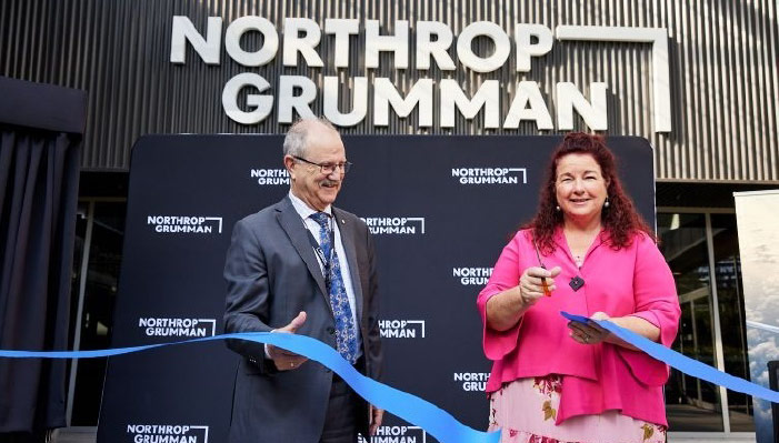 A man and woman stand outside a Northrop Grumman office building and cut a blue ribbon