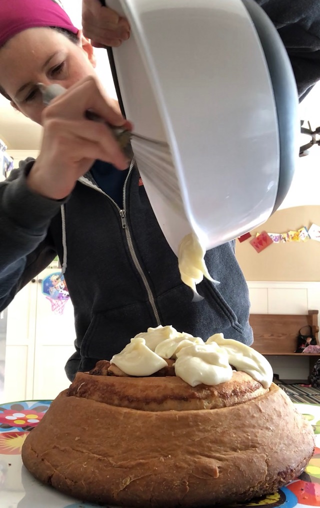 Female holding large mixing bowl, pouring icing on top of huge cinnamon roll.