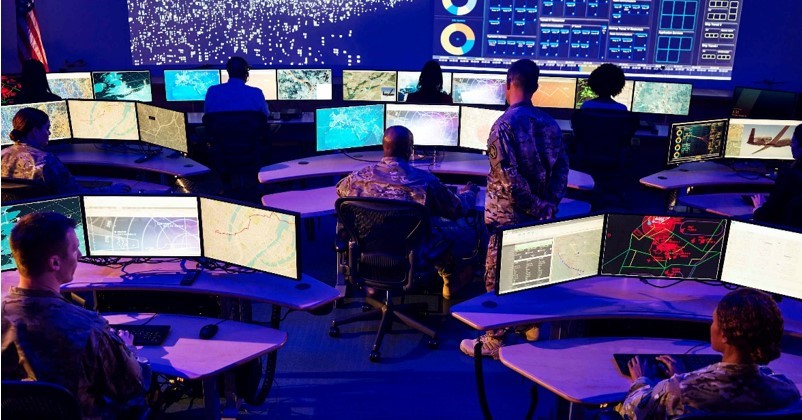 Large command center with multiple computer screens and people monitoring data.